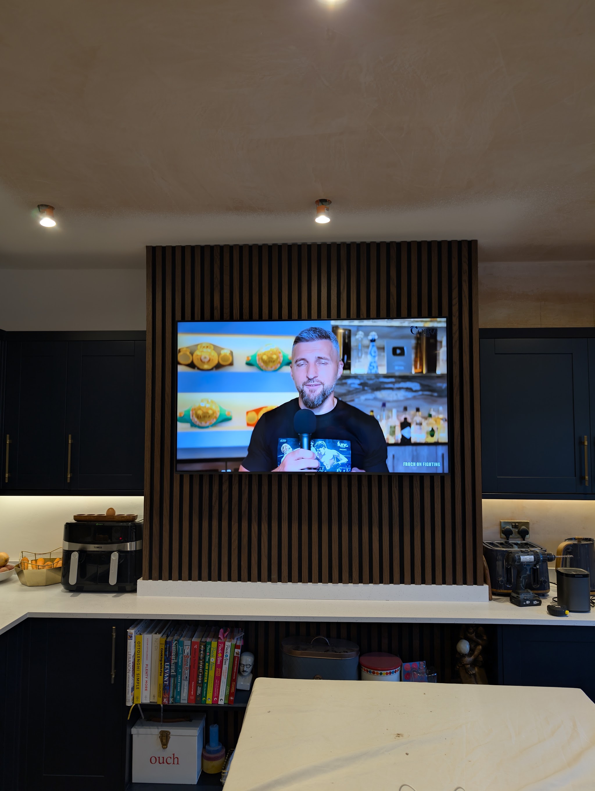 Slatted timber media wall with a mounted TV above navy kitchen units